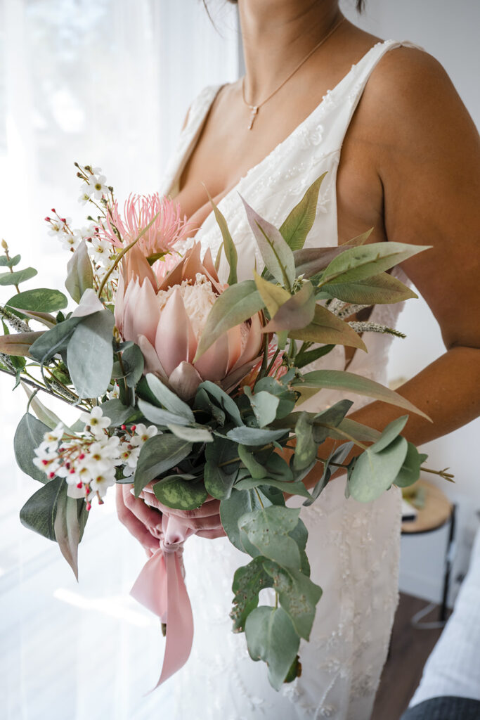 Bride in a white lace dress holding a pink protea bouquet with eucalyptus and white blossoms.