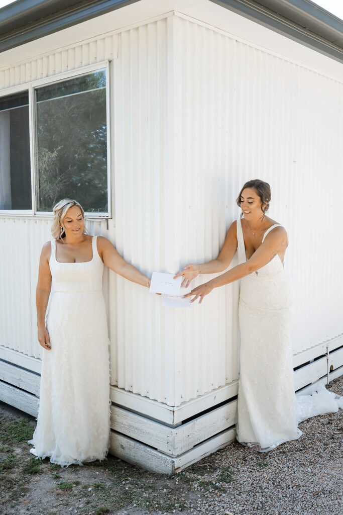 Two brides in wedding dresses exchange envelopes outdoors beside a white corrugated-wall building, smiling.