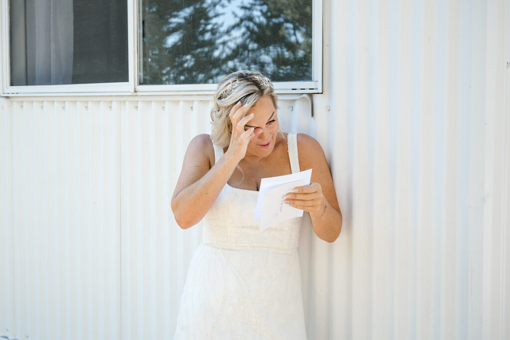 Bride in a white wedding dress reads a letter while leaning against a corrugated metal wall, hand to forehead in emotion.