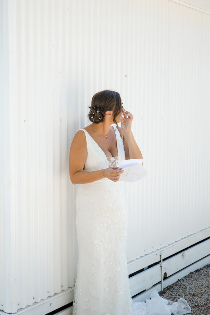 Bride in a lace wedding dress stands beside a white corrugated wall, reading a folded letter.
