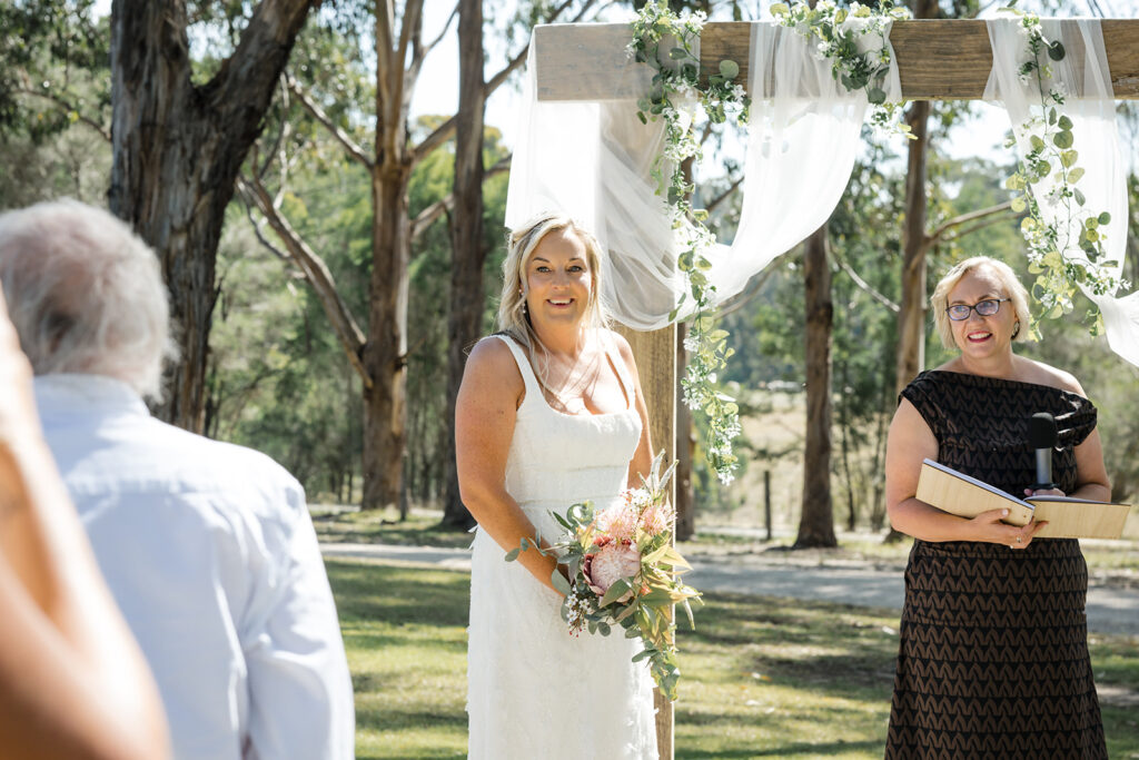 Bride in a white wedding dress holding a bouquet stands under a decorated wooden arch as an officiant speaks to guests outdoors.