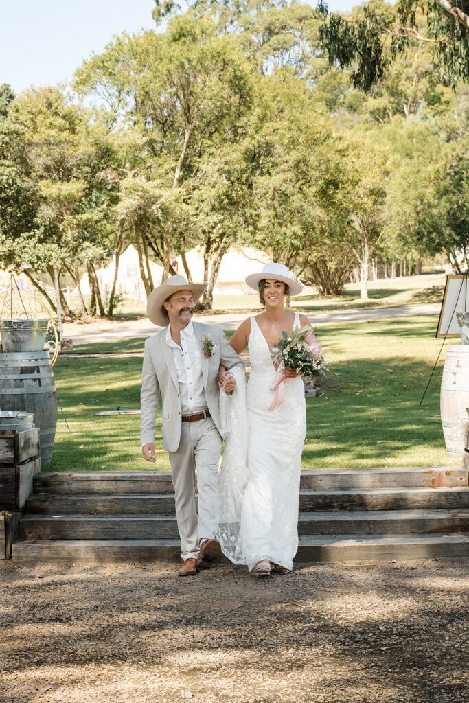 Bride in a white lace wedding dress and a wide-brim hat walks arm-in-arm with a man in a light beige suit and hat outdoors on wooden steps, holding a bouquet and veil.