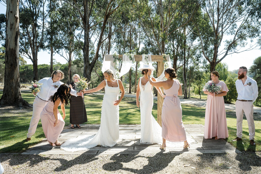 Outdoor wedding scene with two brides in white gowns under a wooden arch, surrounded by bridesmaids in blush pink dresses and guests on a sunlit lawn.