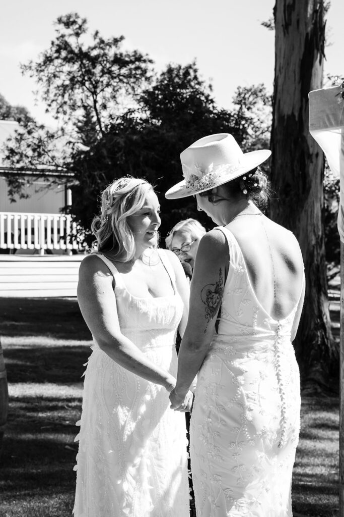 Two brides in wedding dresses hold hands and face each other outdoors, one wearing a hat with flowers.