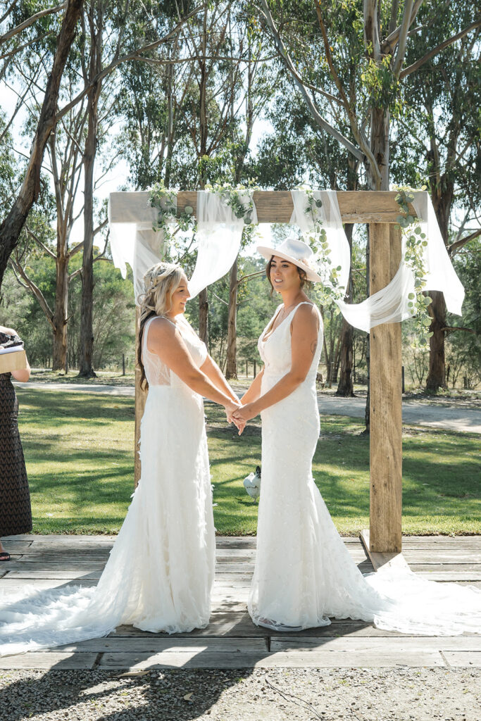 Two brides in white gowns hold hands beneath a wooden arch draped with sheer fabric and greenery in an outdoor setting.