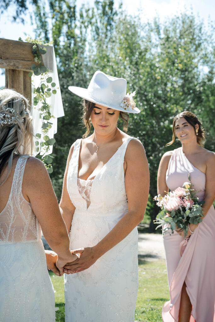 Bride in a white lace dress and white hat stands outdoors, holding hands with another bride as a bridesmaid in a pale pink dress and bouquet looks on.