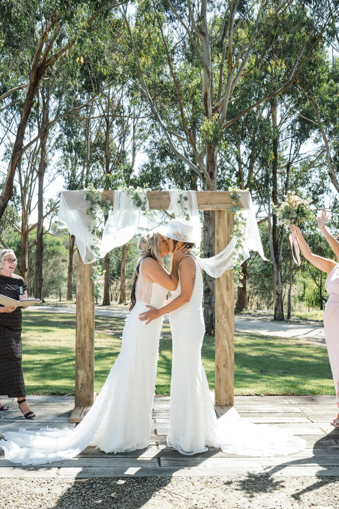Two brides kiss under a wooden, flower-adorned arch during an outdoor wedding ceremony.
