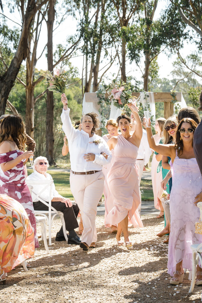 Outdoor wedding celebration with bridesmaids and guests cheering as bouquets are raised in the air along a gravel aisle among trees.