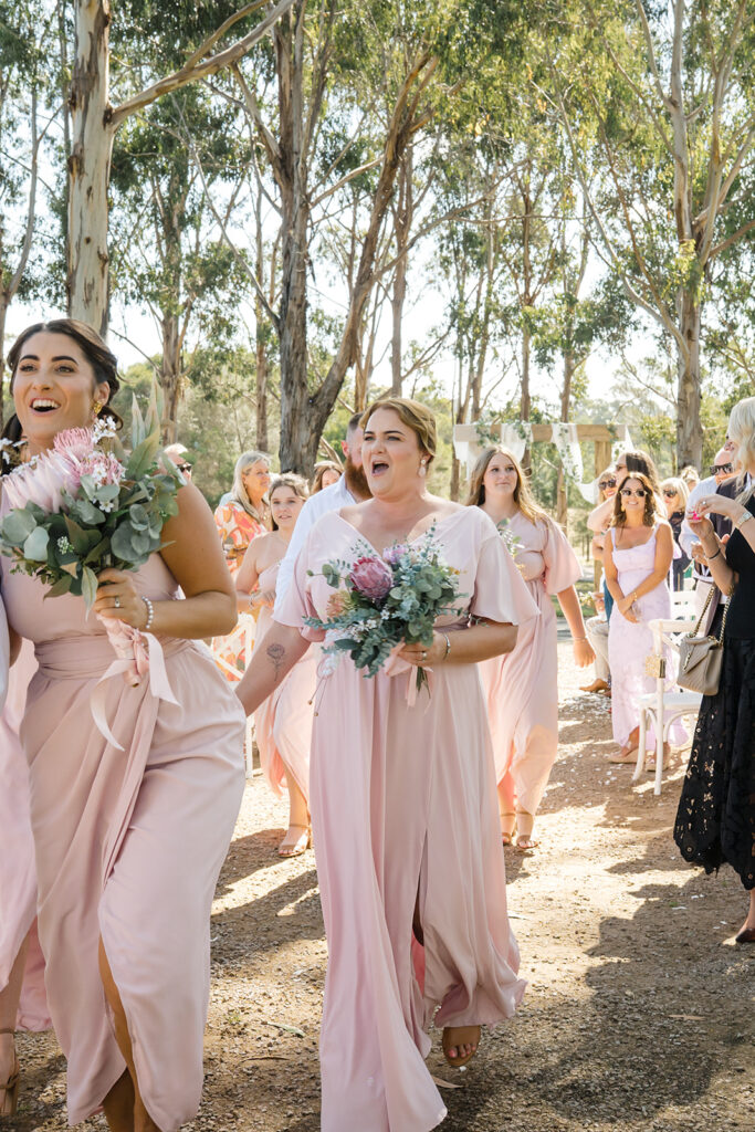 Bridesmaids in pale pink dresses walk down an outdoor ceremony aisle, holding bouquets of pink protea and greenery under tall trees.