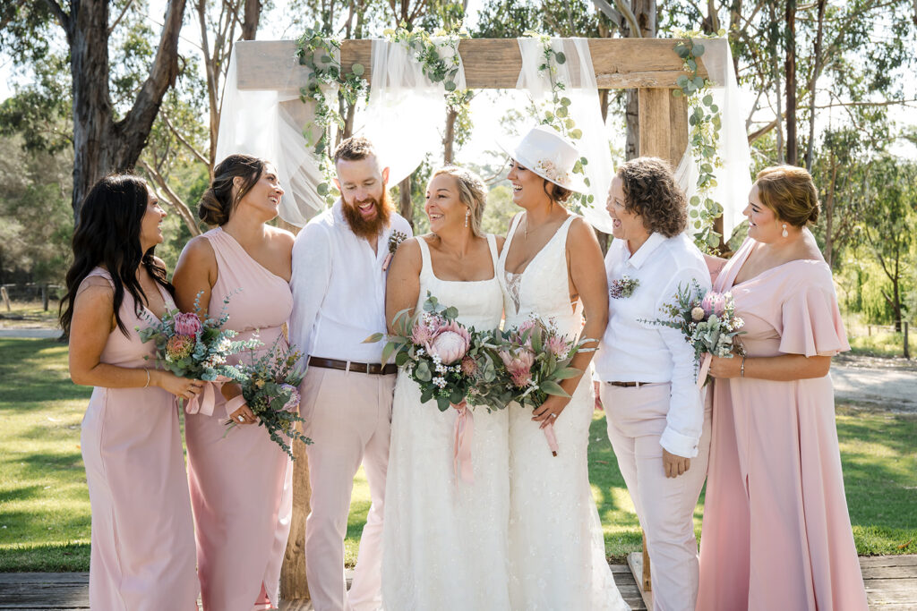 Bride and groom with bridesmaids under a wooden arch decorated with greenery, posing outdoors and smiling with bouquets in pink tones.