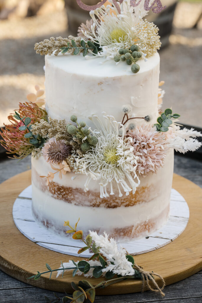 Two-tier naked cake adorned with cream and dusty-pink dried flowers and greens on a wooden board.