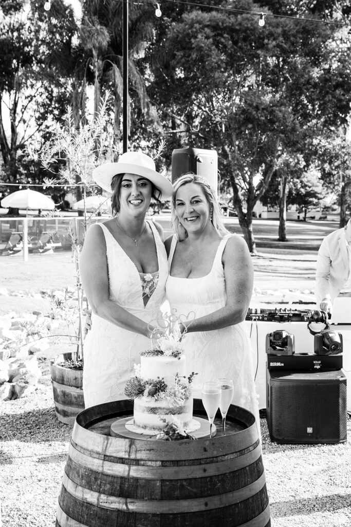 Two women in white dresses smile and cut a wedding cake on a wooden barrel at an outdoor reception, with string lights overhead.