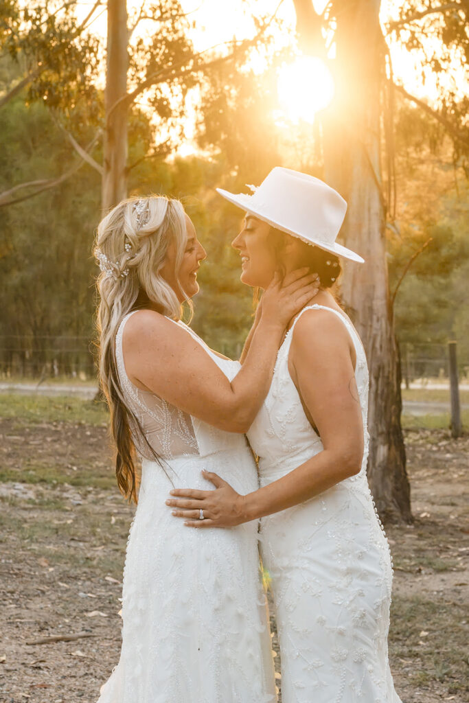 Two brides in white lace dresses embrace each other at sunset in a wooded outdoor setting, with warm backlighting behind them.