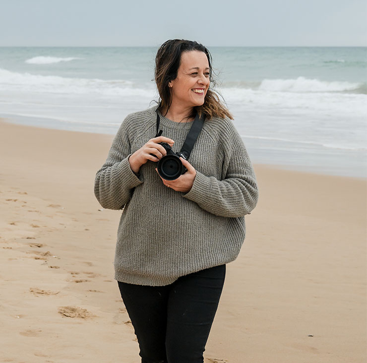 Woman on a sandy beach, smiling, holding a DSLR camera with strap across her chest.