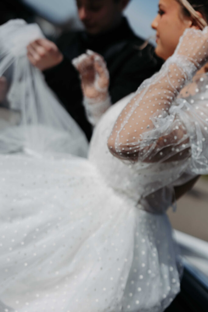 Bride in a white wedding gown with sheer, dotted sleeves adjusting her veil, with a groom in the background.