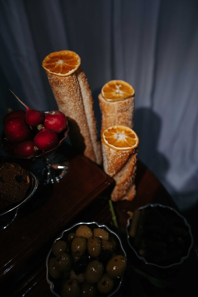 Still-life with crusty bread sticks topped by dried orange slices, beside a bowl of red radishes and bowls of olives on a dark wooden table.