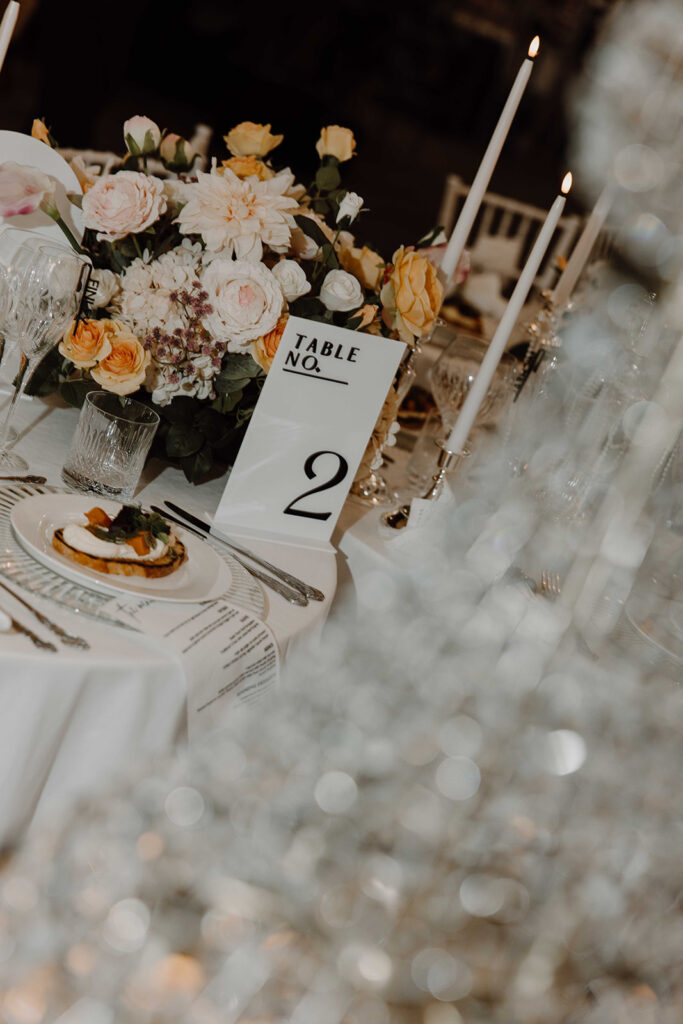 Elegant banquet table with a cream-pink floral centerpiece, tall white candles, glassware, and a 'Table No. 2' place card.