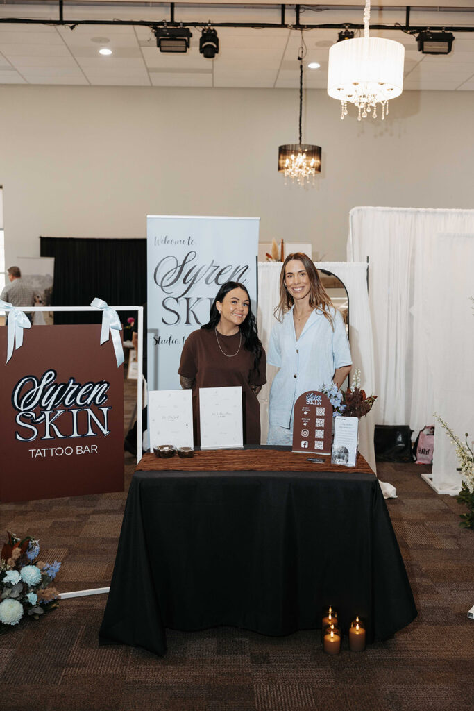 Two women pose at a vendor booth for Syren Skin Tattoo Bar, with banners, a table of brochures, and candles.