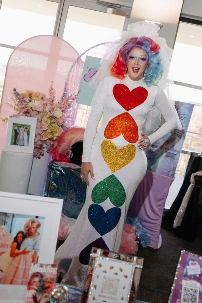 Person wearing a white sequined gown with rainbow heart shapes, colorful wig, and veil posing at a wedding-themed display.