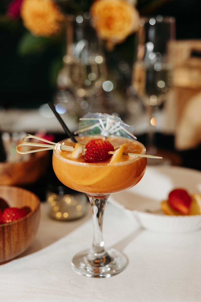 Orange cocktail in a coupe glass with a strawberry and citrus wedge on a skewer, and a mini umbrella, atop a white table.