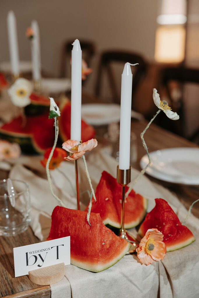 Wedding table centerpiece with tall white candles in gold holders, pastel flowers, and watermelon slices on a linen runner.