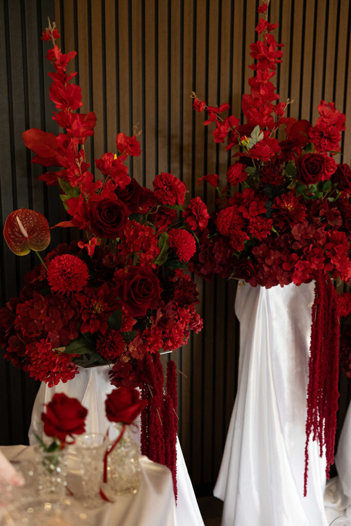 Red flower arrangements with roses and hanging amaranth on white stands against a dark slatted wall, likely decor for a formal event.