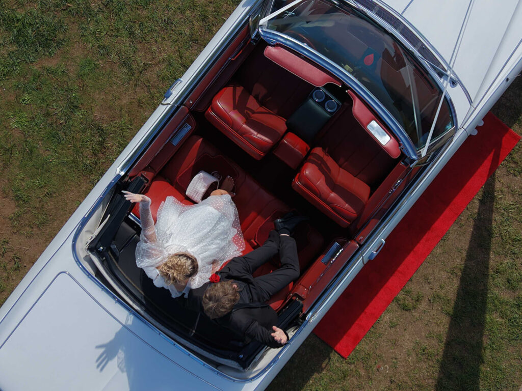 Aerial view of a vintage convertible with red leather interior; bride in a white dress and groom in a black tux seated in the back seat, red carpet outside.