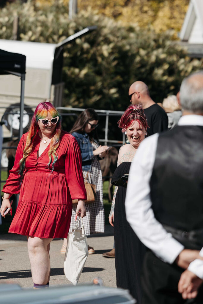 Two women at an outdoor event; one in a bright red dress with rainbow hair and heart-shaped sunglasses smiling beside another woman with pink hair and tattoos.