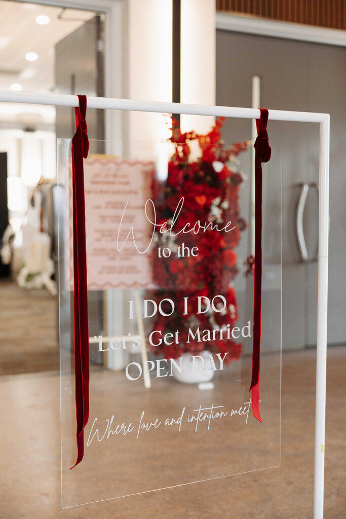 Transparent acrylic sign on a white frame with red ribbons, reading 'Welcome to the I DO I DO Let's Get Married OPEN DAY' with a red floral arrangement in the background.