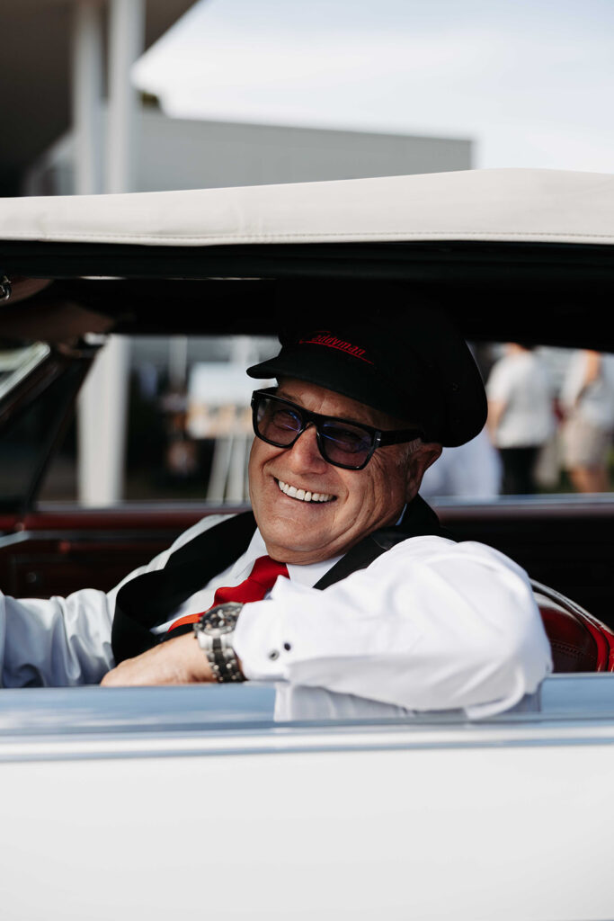 Smiling man in a black cap and glasses sits in a vintage convertible with the top down, wearing a white shirt and red tie