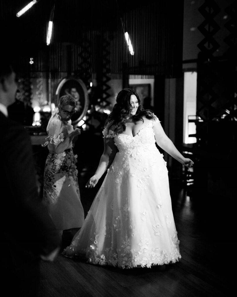 rick_liston1 - I do I do Gippsland Bride in a lace wedding gown dancing on a reception floor, smiling with guests in the dim background (black and white).