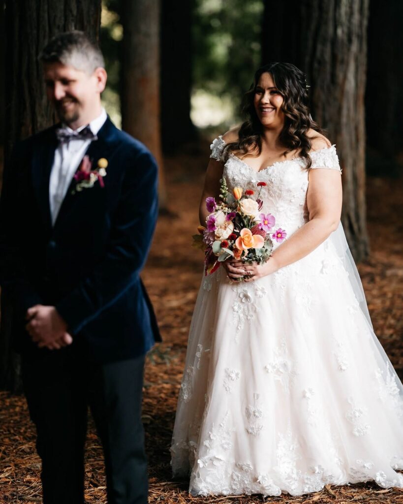 rick_liston3 - I do I do Gippsland Bride in a white lace wedding gown holding a colorful bouquet, standing among trees with the groom blurred beside her in a suit.