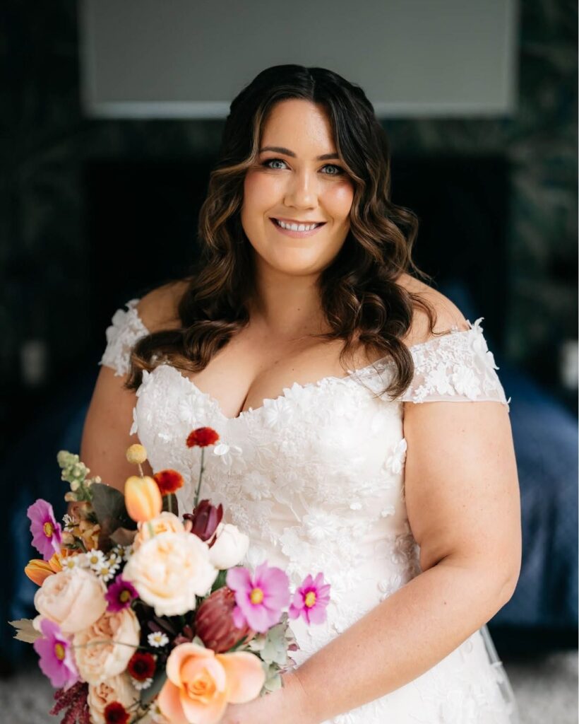 rick_liston4 - I do I do Gippsland Bride in a white lace off-shoulder wedding dress smiling, holding a colorful bouquet of flowers in front of her.