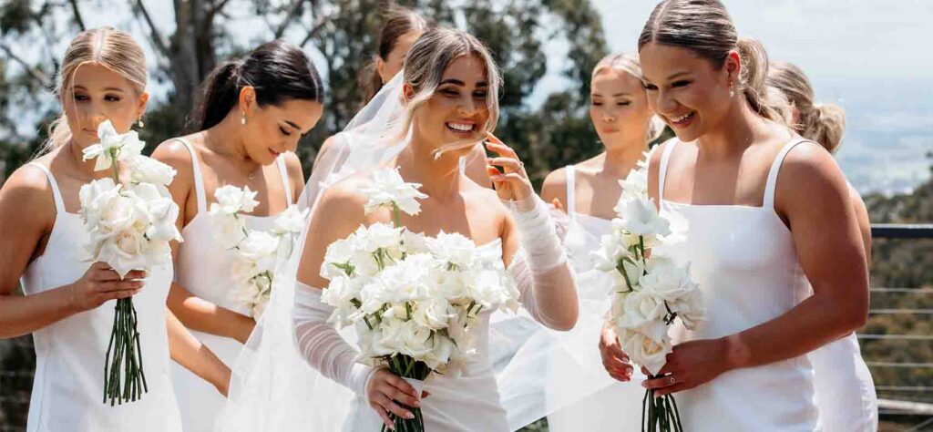 | I do I do Gippsland Bride in white gown with veil, surrounded by bridesmaids in white dresses holding white bouquets outdoors.