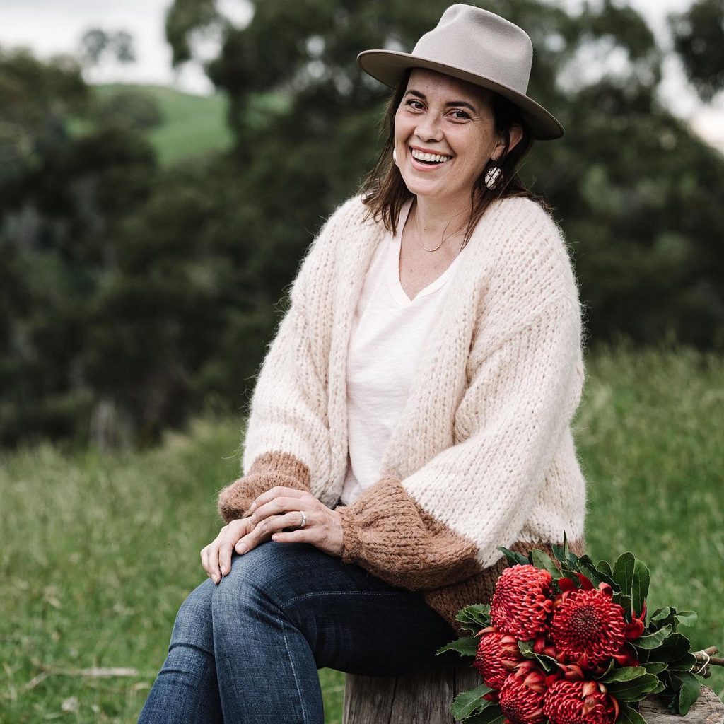 470183263_1292950245388748_970847589132211980_n | I do I do Gippsland Smiling woman in a beige hat and cream cardigan sits outdoors on a log, with a red bouquet beside her on the ground.