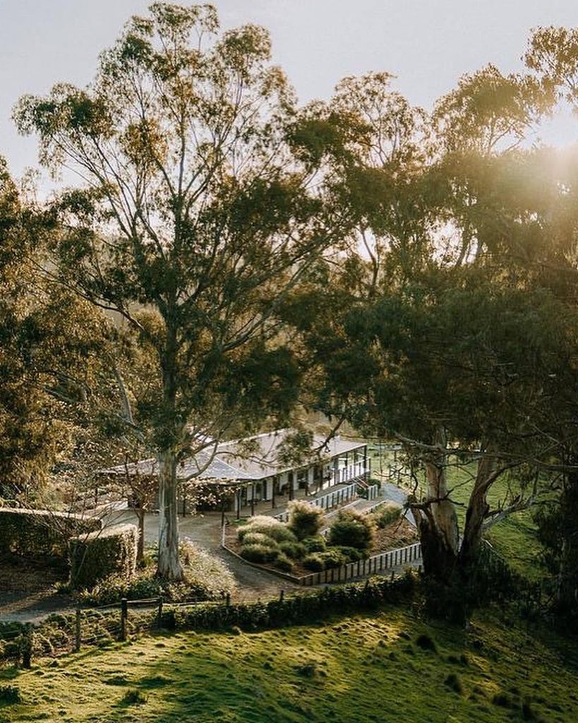 471962082_1305079574175815_189746001003097394_n 2 | I do I do Gippsland Sunlit park scene with large trees, a courtyard building with a wraparound covered walkway, and trimmed shrubs below.