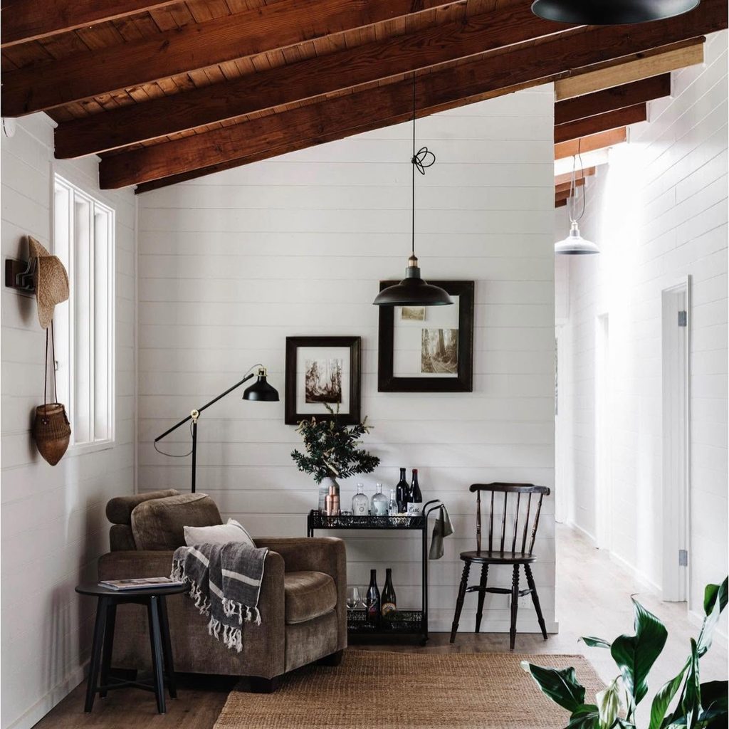475492521_1181724303700611_5443432899821000621_n | I do I do Gippsland Cozy living room with white paneled walls, dark wooden beams, and a brown sofa near a black bar cart. A pendant light hangs from the ceiling.