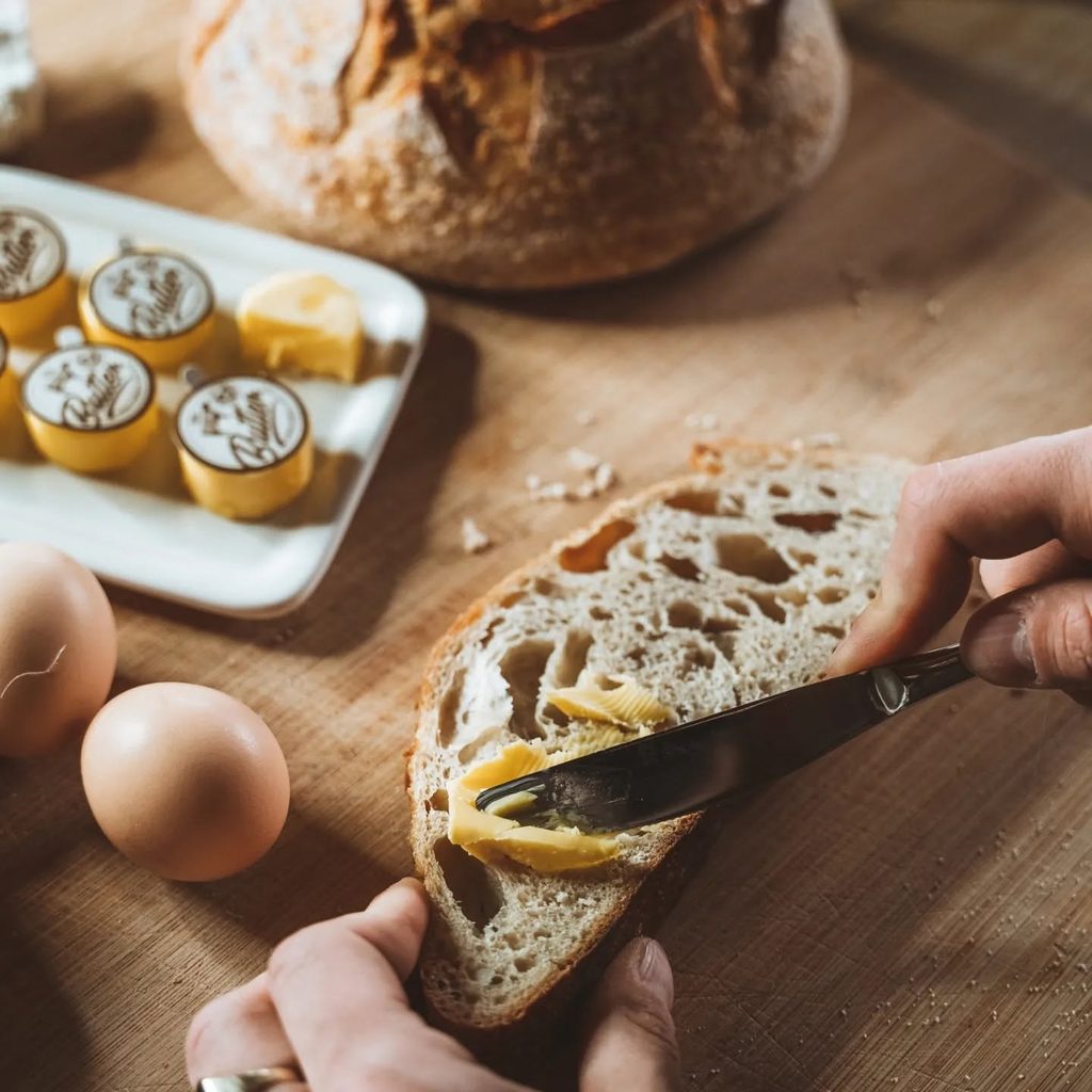 488234156_1231621582044216_8126670673933146369_n | I do I do Gippsland Hands spread butter on a slice of rustic bread with a knife, in a warm kitchen scene.