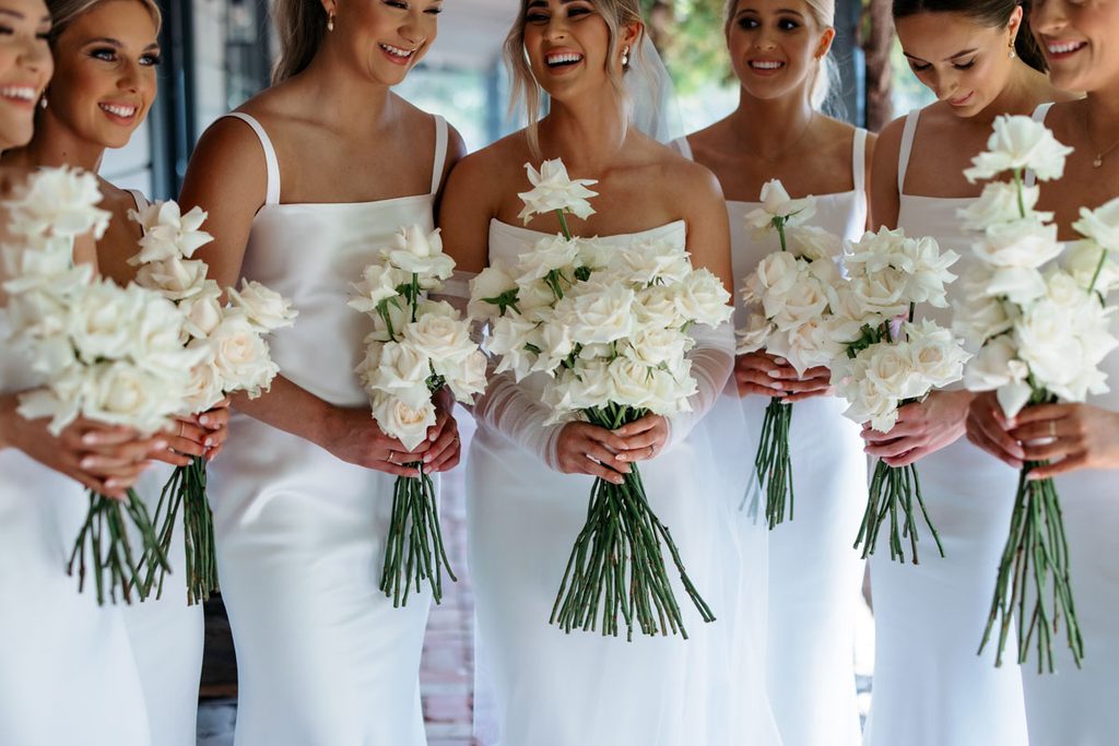 Mia + Thomas | I do I do Gippsland Bridesmaids in white dresses holding white bouquet stems, smiles visible as a group portrait.