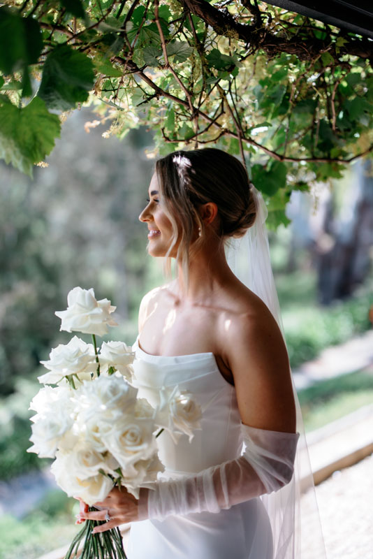 Mia + Thomas | I do I do Gippsland Bride in a strapless white wedding dress under greenery, holding a bouquet of white roses and wearing a veil.