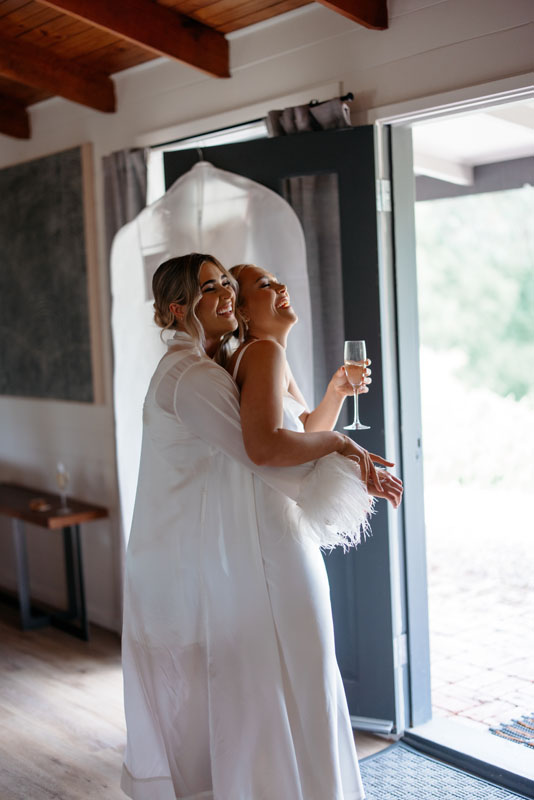 Mia + Thomas | I do I do Gippsland Two brides in white robes embracing and laughing, one holding a champagne flute by an open doorway inside a sunlit room.