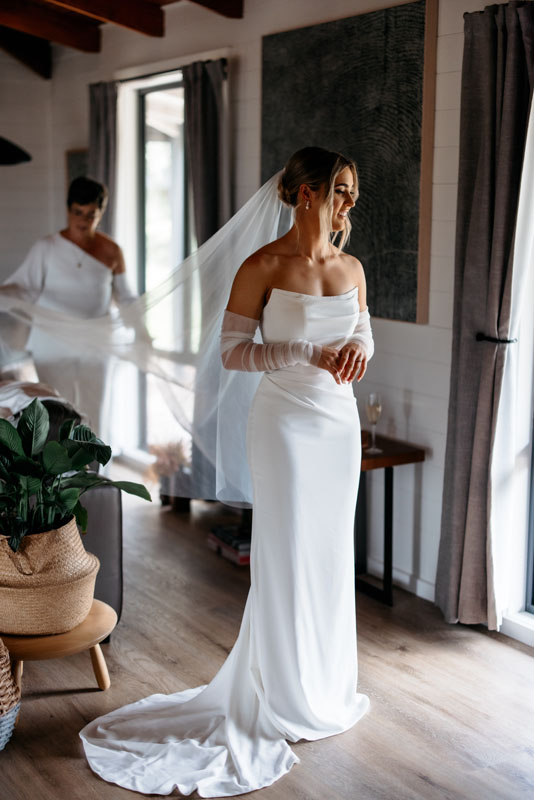 Mia + Thomas | I do I do Gippsland Bride in a white off-the-shoulder gown with a long veil, standing by a window as a second guest adjusts her veil in the background.