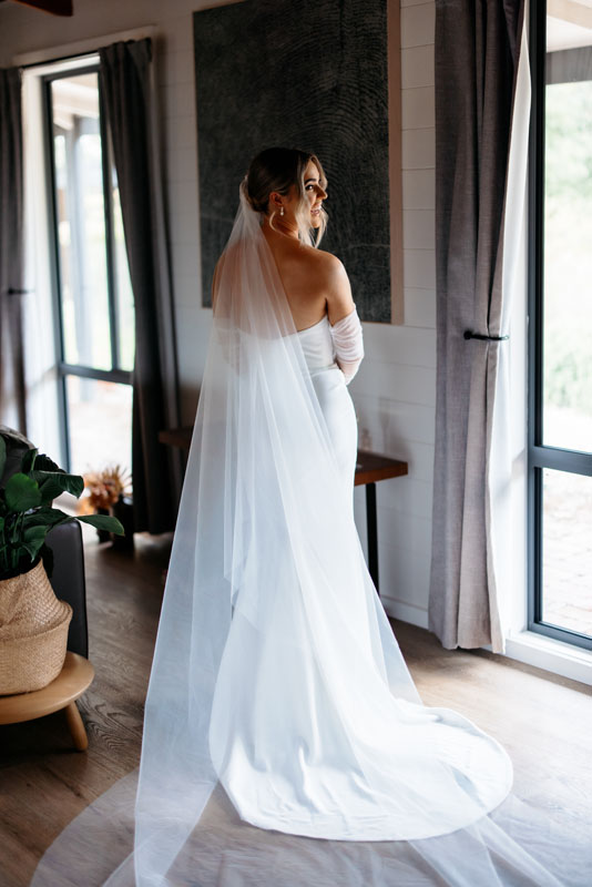 Mia + Thomas | I do I do Gippsland Bridal gown with long veil, viewed from behind, standing indoors near windows and curtains.
