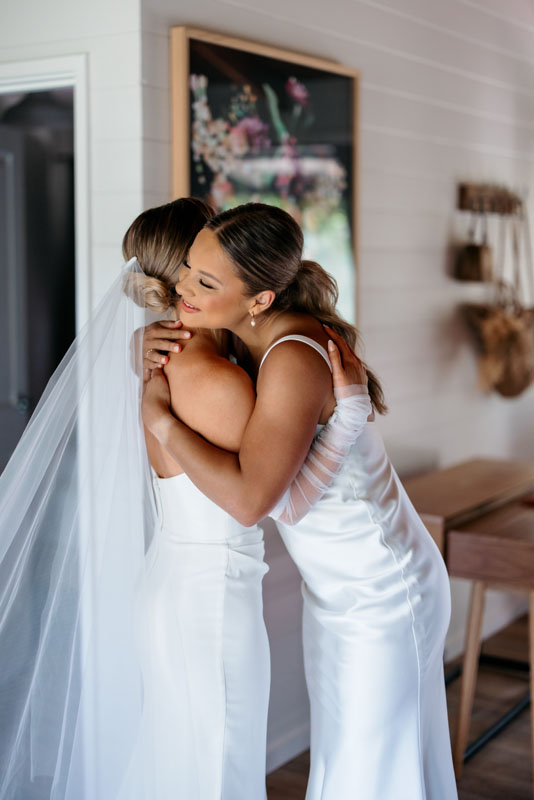 Mia + Thomas | I do I do Gippsland Two brides in white wedding gowns embrace in a cozy room, one with a veil trailing behind her records the moment of joy.