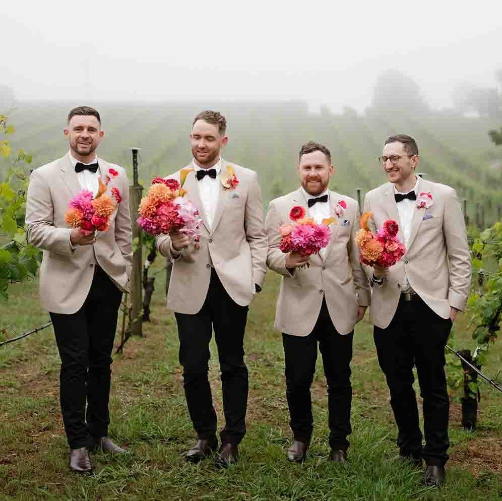1 | I do I do Gippsland Four groomsmen in beige blazers and black bow ties stand in a foggy vineyard, each holding bright pink and orange bouquets at chest level in a casual pose.