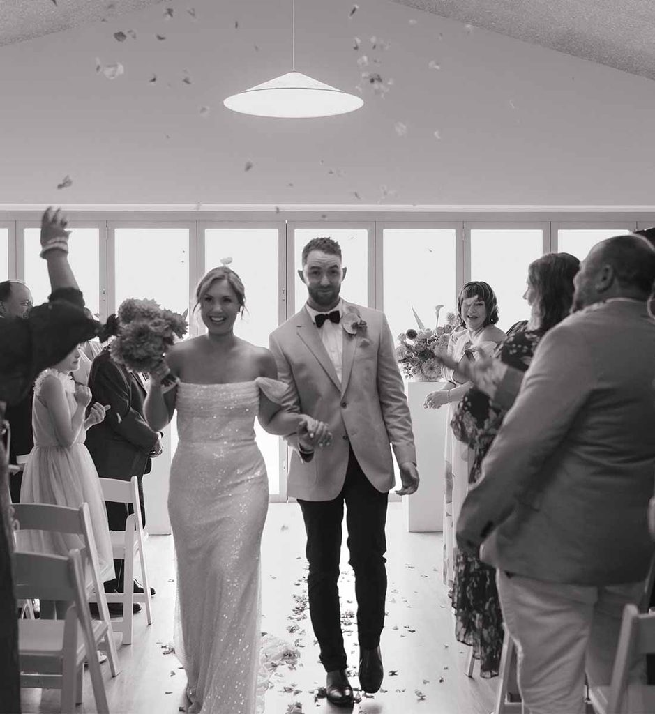 2 | I do I do Gippsland Bride and groom walk arm in arm down the aisle as guests celebrate, confetti in the air (black-and-white photo).