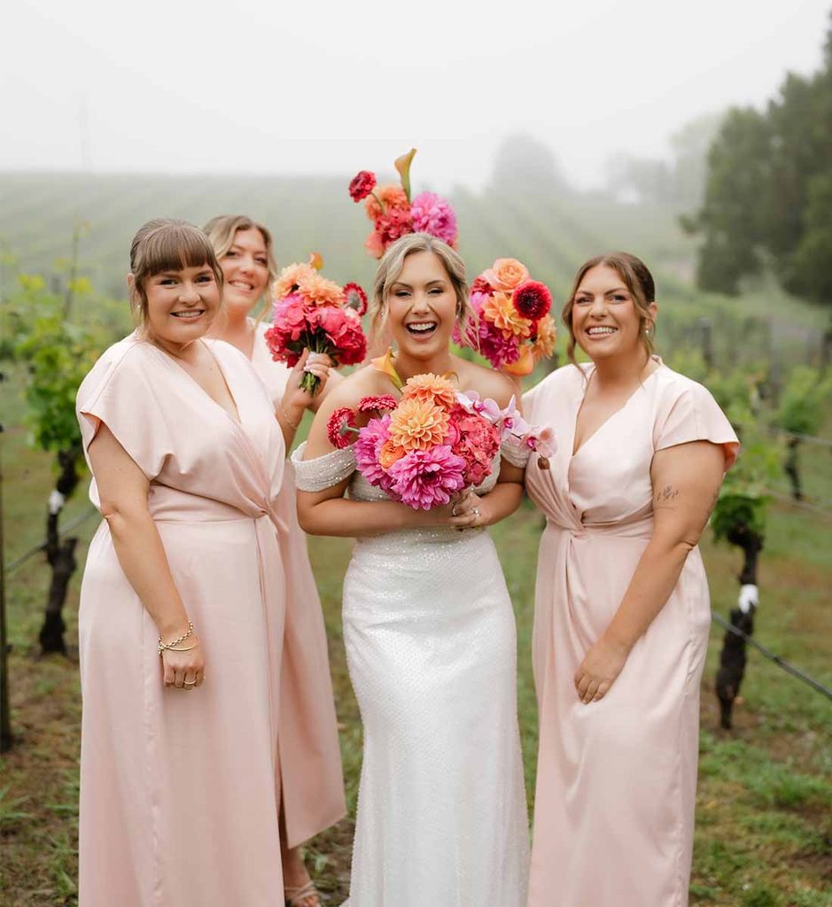 3 | I do I do Gippsland Bride in a white gown holding a colorful bouquet, posing with four bridesmaids in blush dresses in a vineyard.