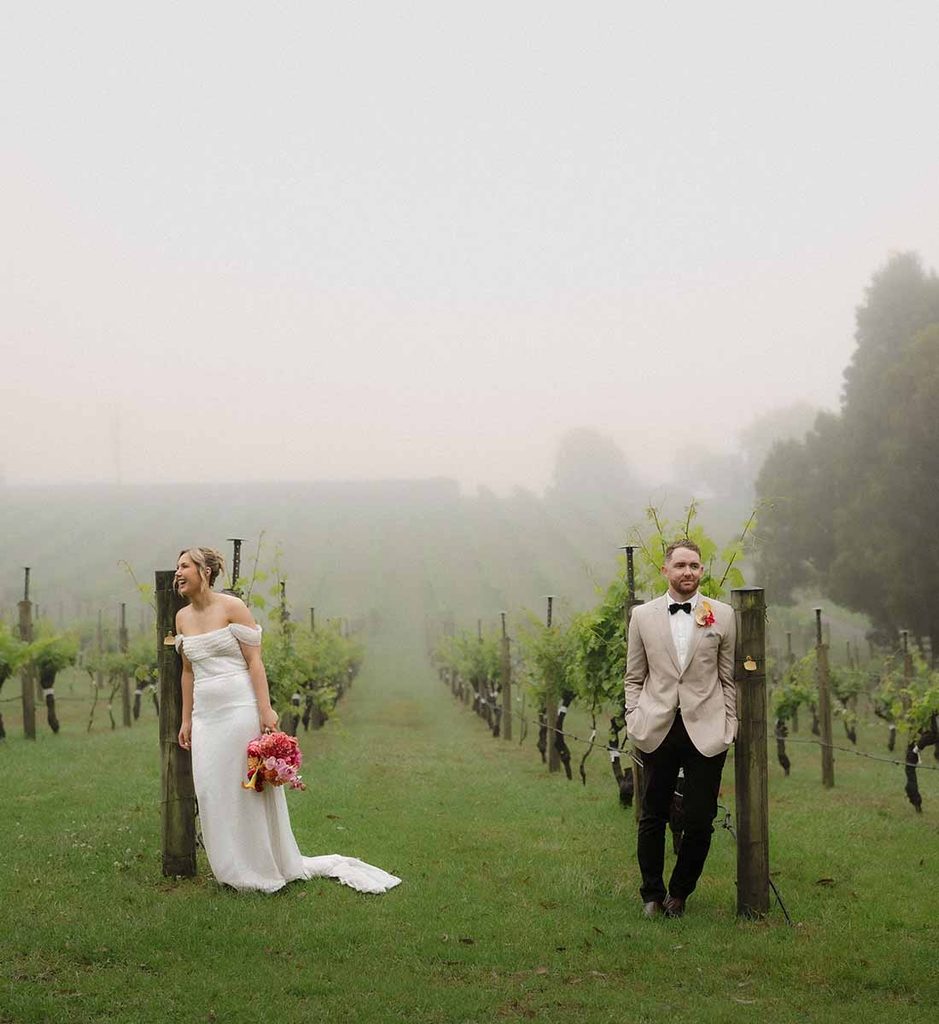 4 | I do I do Gippsland Bride in a white off-shoulder gown holds a pink bouquet on a foggy vineyard; groom in a beige suit stands nearby.