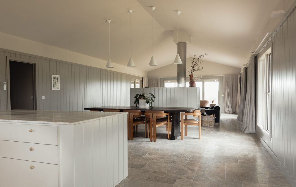 CarrajungEstate_Preview_2026-013 | I do I do Gippsland Open-plan kitchen and dining area with grey paneled walls, five white pendant lights above a long wooden table, and a central column.