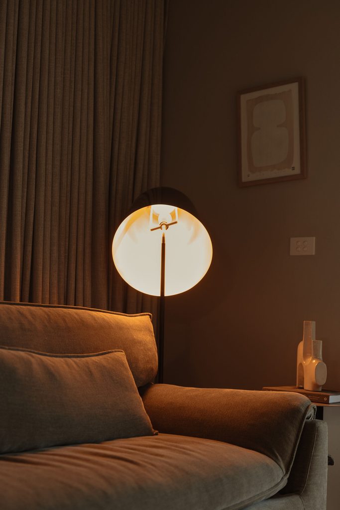 CarrajungEstate_Preview_2026-051 | I do I do Gippsland Dimly lit living room corner with a beige sofa, a glowing floor lamp, heavy brown curtains, and a framed abstract painting on the wall.